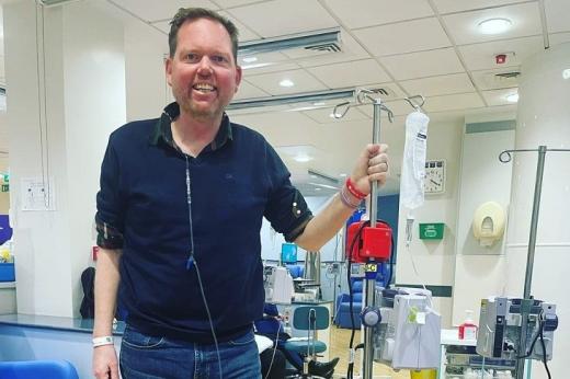 A patient smiling and standing in a Royal Marsden hospital room, attached to a drip