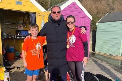 Bowel cancer patient Ben, standing with his arm around his friend and young godson, who are wearing Royal Marsden Cancer Charity t-shirts and medals. They are standing in front of some colourful beach huts on a sunny day.